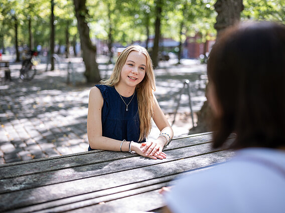 Eine junge Frau sitzt in einem Park an einem Holztisch und unterhält sich.