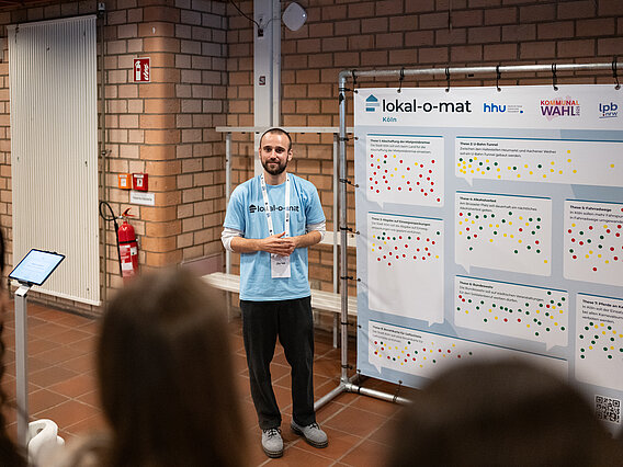 Ein junger Mann steht neben einer großen Stellwand. Er trägt ein T-shirt mit dem Aufdruck "lokal-o-mat".