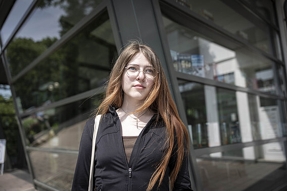 Eine junge Frau steht vor einer Fensterfront.