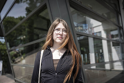 Eine junge Frau steht vor einer Fensterfront.