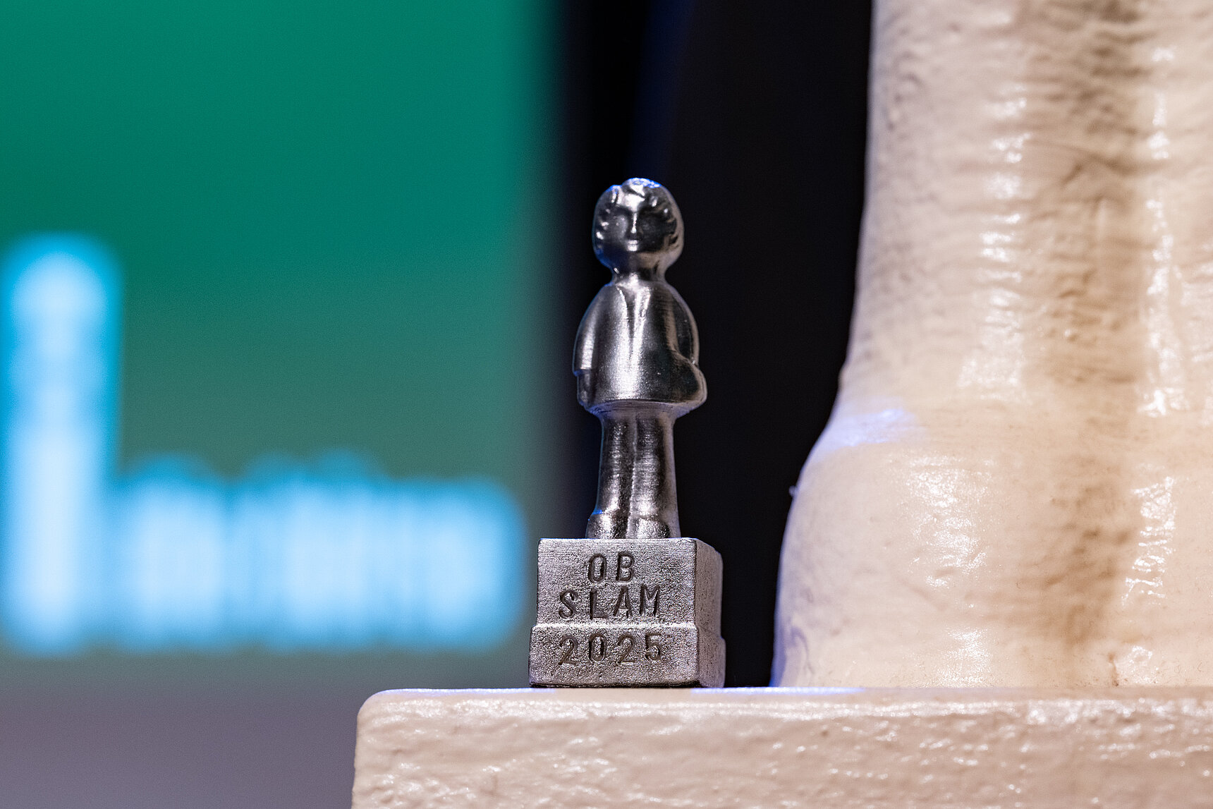 Das Bild zeigt die Statue zum Kandidatenslam in Wuppertal: eine kleine silberne Figur, auf deren Sockel OB-Slam 2025 steht.